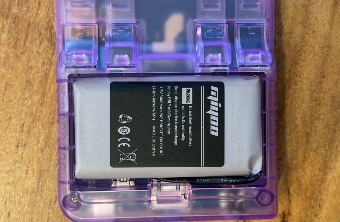 Intérieur du Miyoo Mini Plus montrant la batterie Li-ion 3,7V 300mAh, avec coque violette transparente ouverte sur une surface en bois.